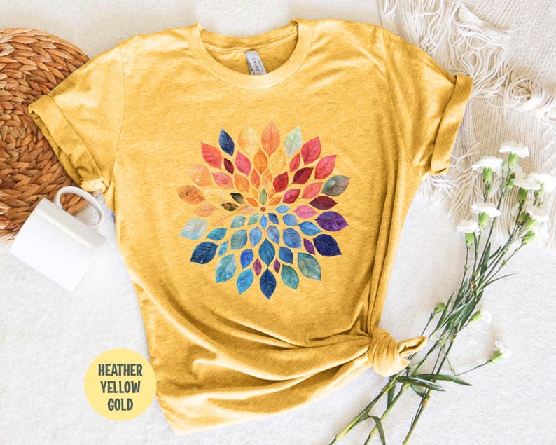 K&ouml;nnte beinhalten: Ein heather-gelbgoldenes T-Shirt mit einem kreisf&ouml;rmigen Grafikdesign, das aus aquarellartigen Bl&auml;ttern in einem Farbspektrum aus Rot, Orange, Gelb, Gr&uuml;n, Blau und Violett besteht, die in einem radialen Muster auf der Brust angeordnet sind.