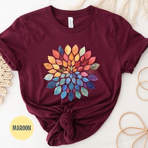 K&ouml;nnte beinhalten: Ein kastanienbraunes Kurzarm-T-Shirt mit einem kreisf&ouml;rmigen Grafikdesign aus bunten Bl&auml;ttern, die in einem radialen Muster angeordnet sind. Die Bl&auml;tter zeigen ein Farbspektrum von Gelb, Orange, Rot, Pink, Violett bis hin zu Blau.