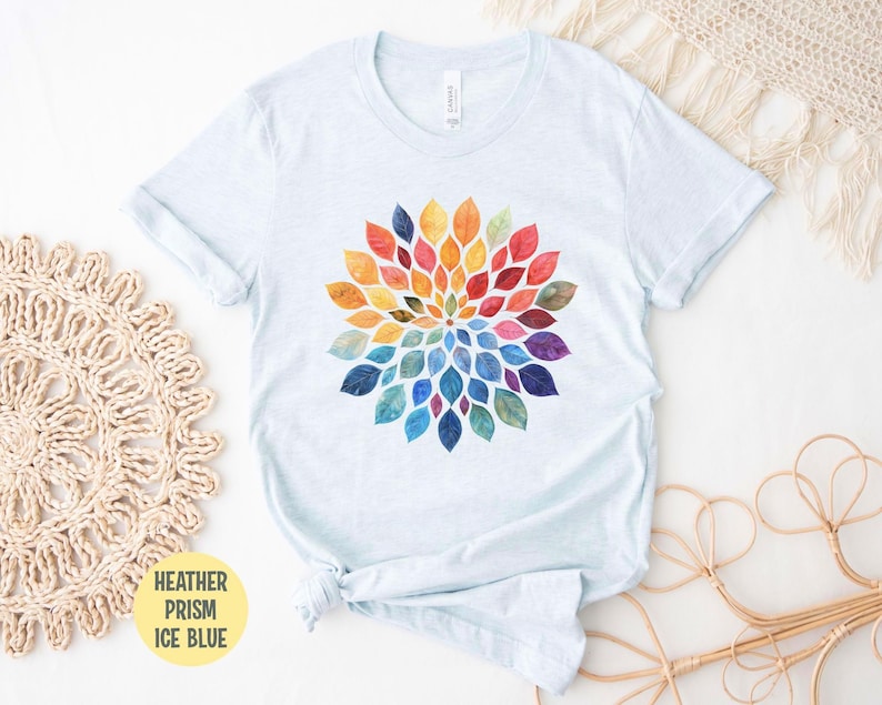 K&ouml;nnte beinhalten: Ein eisblaues, meliertes T-Shirt mit einer zentralen kreisf&ouml;rmigen Grafik aus aquarellartigen Bl&auml;ttern, die in einem lebendigen Regenbogenspektrum angeordnet sind und von einem kleinen Mittelpunkt aus strahlenf&ouml;rmig verlaufen.