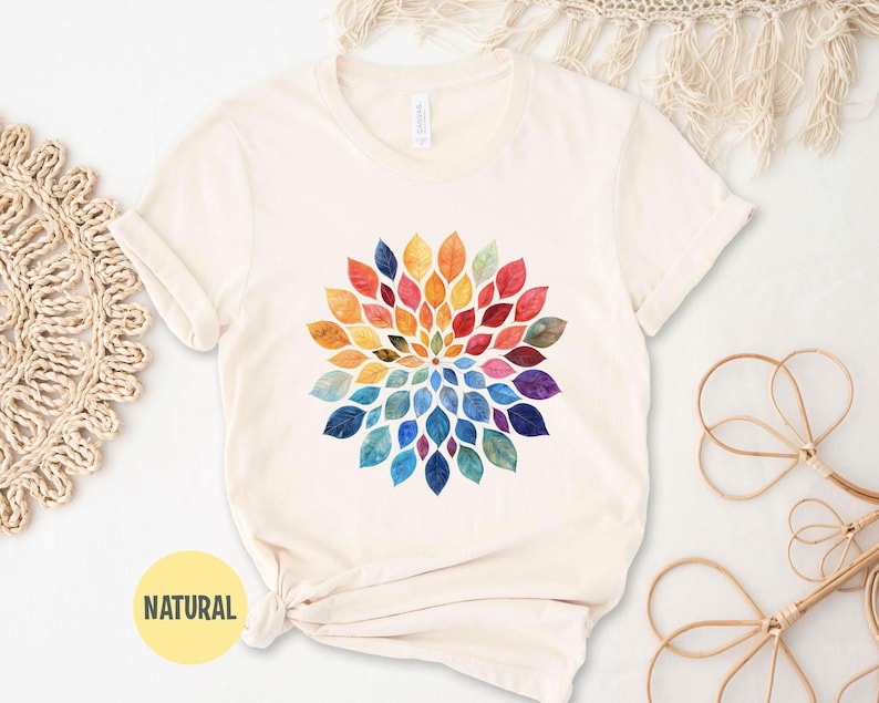 K&ouml;nnte beinhalten: Ein cremefarbenes Kurzarm-T-Shirt mit einer kreisf&ouml;rmigen Mandala-Grafik, die aus aquarellartigen Bl&auml;ttern in einem Farbspektrum aus Rot, Orange, Gelb, Gr&uuml;n, Blau und Violett besteht, welche in einem radialen Muster angeordnet sind.