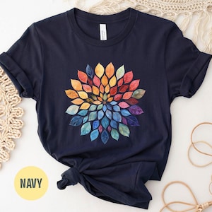 K&ouml;nnte beinhalten: Ein marineblaues Kurzarm-T-Shirt mit einer kreisf&ouml;rmigen Mandala-Grafik, die aus aquarellartigen Bl&auml;ttern in einem Farbspektrum aus Gelb, Orange, Rot, Violett und Blau besteht und in einem radialen Muster auf der Brust angeordnet ist.