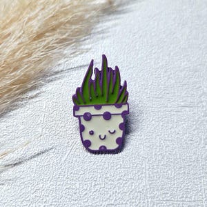 Könnte beinhalten: Ein skurriler Emaille-Pin mit einem Topfpflanzendesign. Der Pin zeigt einen weißen Topf mit lila Punkten und einem lächelnden Gesicht. Die Pflanze hat grüne Blätter und lila Spitzen, perfekt für Pflanzenliebhaber.