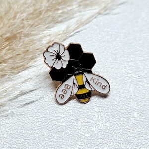 Könnte beinhalten: Goldfarbene Emaille-Anstecknadel mit einem Bienen-Design und dem Schriftzug "Bee Kind". Die Anstecknadel hat eine weiße Blume und eine schwarze Wabenform. Die Biene hat gelbe und schwarze Streifen.