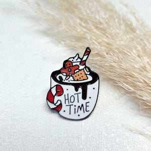 Könnte beinhalten: Weißer Emaille-Pin, der eine Tasse heiße Schokolade mit Schlagsahne, einem Lebkuchenmann und einer Zuckerstange darstellt. Die Tasse hat einen rot-weißen Zuckerstangen-Griff und die Worte "HOT TIME".