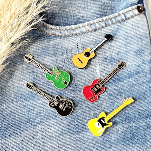 Könnte beinhalten: Fünf gitarrenförmige Emaille-Anstecknadeln in verschiedenen Farben auf blauem Denim-Hintergrund. Die Anstecknadeln umfassen grüne, schwarze, rote und zwei gelbe Gitarren. Die Anstecknadeln haben schwarze Umrisse und silberne Saiten.