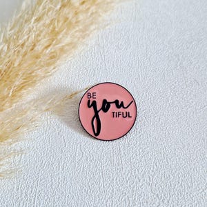Könnte beinhalten: Runder, rosa Emaille-Ansteckpin mit den Worten "BE you TIFUL" in schwarzer Schrift. Der Pin hat einen schwarzen Rand und ist vor einem strukturierten weißen Hintergrund platziert. Getrocknetes Gras ist im Hintergrund zu sehen.