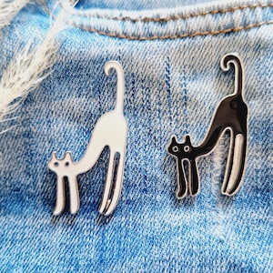 Könnte beinhalten: Zwei Emaillestifte mit Katzen mit langen Beinen. Eine Katze ist schwarz und die andere ist weiß. Die Stifte befinden sich auf einem blauen Jeansstoff.