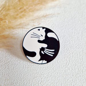 Könnte beinhalten: Runder Ansteckpin mit einem Yin-Yang-Design aus zwei Katzen, eine weiß und eine schwarz. Der Pin befindet sich auf einem strukturierten weißen Hintergrund, mit einer verschwommenen beigen Pflanze im Hintergrund. Katzen-Accessoire.