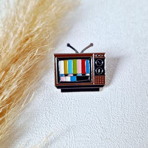 Könnte beinhalten: Ein Emaille-Pin im Retro-Stil in Form eines Vintage-Fernsehers. Der Fernseher hat einen braunen Rahmen, einen Bildschirm mit bunten Testbalken und eine silberne Antenne. Der Pin ist ein kleines, dekoratives Accessoire.