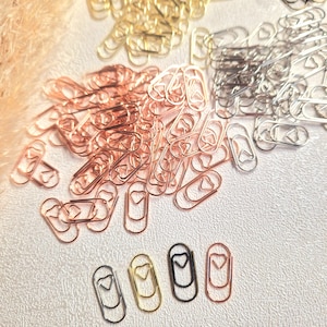 Könnte beinhalten: Ein großer Haufen herzförmiger Büroklammern in Gold, Roségold und Silber. Vier einzelne Klammern sind in einer Reihe unter dem Haufen angeordnet, eine in Gold, eine in Roségold, eine in Silber und eine in Schwarz.