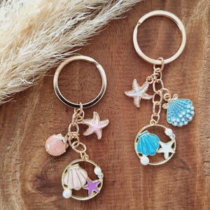 Könnte beinhalten: Zwei goldene Schlüsselanhänger mit Muschel- und Seestern-Motiv. Ein Schlüsselanhänger hat eine blaue Muschel, einen weißen Seestern und eine rosa Muschel. Der andere Schlüsselanhänger hat eine rosa Muschel, einen lila Seestern und eine weiße Perle.