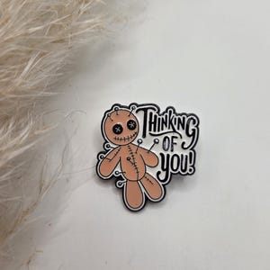 Könnte beinhalten: Hellbraune Voodoo-Puppen-Anstecknadel mit schwarzen Umrissen und dem Text "Thinking of you!". Die Puppe hat schwarze kreuzförmige Augen und ist zusammengenäht. Die Anstecknadel befindet sich auf einer weißen Oberfläche.