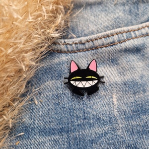 Könnte beinhalten: Schwarze Emaille-Anstecknadel mit einem Cartoon-Katzenmotiv, das ein breites, zahniges Grinsen zeigt. Die Katze hat rosa Ohren und gelbe Zähne. Die Anstecknadel befindet sich auf einem blauen Jeansstoff.