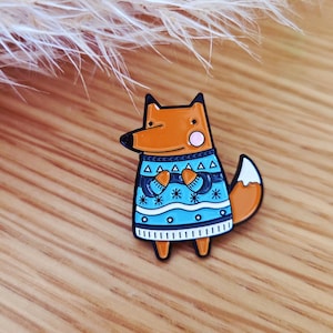 Könnte beinhalten: Ein farbenfroher Emaille-Pin mit einem Cartoon-Fuchs, der einen blauen Pullover mit Schneeflocken- und geometrischen Mustern trägt. Der Fuchs hat orangefarbenes Fell, einen weißspitzigen Schwanz und schwarze Akzente.