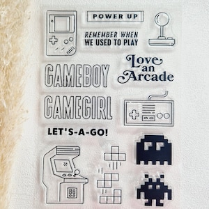 Könnte beinhalten: Klares Stempelset mit Retro-Videospiel-Designs. Enthält einen Gameboy, eine Spielhalle, einen Controller und pixelige Charaktere. Der Text enthält "Power Up", "Remember When We Used to Play", "Gameboy", "Gamegirl", "Let's-a-go!" und "Love an Arcade."