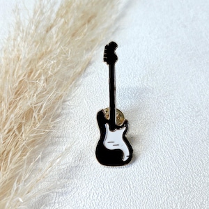 Könnte beinhalten: Ein schwarz-weißes Gitarren-Emaille-Anstecknadel. Der Anstecker zeigt ein klassisches E-Gitarren-Design mit schwarzem Korpus, weißem Schlagbrett und schwarzem Hals. Ein goldfarbener Nadelrücken ist sichtbar.