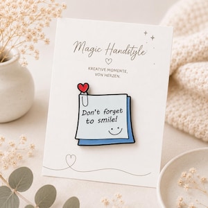 Könnte beinhalten: Eine weiße Emaille-Anstecknadel in Form eines quadratischen Haftnotizzettels mit blauem Rand, einer schwarzen Büroklammer und einem kleinen roten Herz. Die Nadel zeigt den schwarzen Text Don't forget to smile! und ein einfaches schwarzes Smiley-Gesicht.