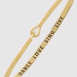 May include: A gold-toned metal bangle bracelet featuring a hook-and-eye style closure mechanism. The outer surface of the thin, polished band is engraved with the black text DANCE LOVE SING LIVE, positioned clearly along the length of the metal.