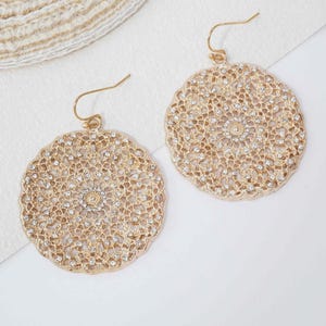 May include: A pair of gold-toned dangle earrings featuring a circular, intricate filigree design. Each earring is embellished with small, clear sparkling stones embedded throughout the metal pattern and hangs from a simple gold-toned hook wire.