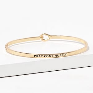 May include: A gold-toned metal bangle bracelet featuring a smooth, polished finish and a teardrop-shaped clasp mechanism. The outer surface of the bracelet is engraved with the black text "PRAY CONTINUALLY" in a clean, sans-serif font.