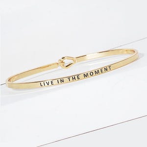 May include: A gold-toned metal bangle bracelet featuring a smooth, polished finish and a central knot-style clasp. The outer surface of the bracelet is engraved with the black text "LIVE IN THE MOMENT" in a clean, sans-serif capital letter font.