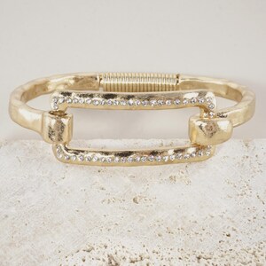 May include: A gold-toned metal hinged bracelet featuring a rectangular center link adorned with small, clear, sparkling crystals. The bracelet has a textured, matte finish and a sturdy, wide band design that secures with a spring-loaded hinge mechanism.