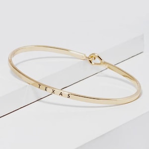 May include: A gold-toned metal bangle bracelet featuring a smooth, polished finish. The word TEXAS is engraved in black capital letters along the front of the band. The bracelet has a simple hook-and-eye style closure mechanism on one side.