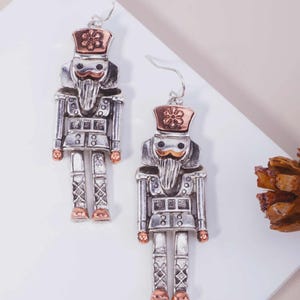 May include: A pair of silver-toned nutcracker earrings with copper-colored accents on the hat, mustache, hands, and feet. Each earring features a detailed soldier figure with a tall hat, a long beard, and a uniform, suspended from a silver hook.