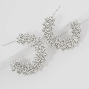 May include: A pair of silver-toned hoop earrings featuring a complex, three-dimensional geometric lattice structure. Each earring is constructed from a repeating pattern of small, interconnected pyramid shapes, creating a textured, openwork design.