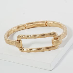 May include: A gold-toned metal bracelet featuring a textured, hammered surface. The design includes a central rectangular link connected to a curved band, which is finished with a coiled spring hinge mechanism for closure and flexibility.