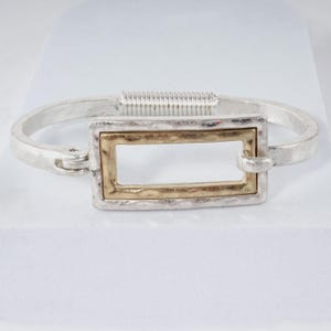 May include: A silver-toned hinged bangle bracelet featuring a central rectangular element with a hammered gold-toned finish. The bracelet has a textured, metallic surface and a secure hinge mechanism on one side, showcasing a two-tone design.