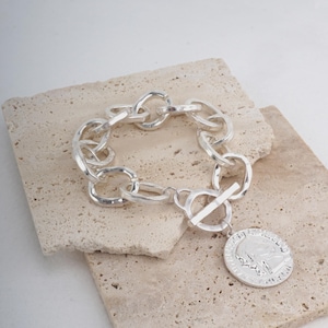 May include: A silver-toned chain link bracelet featuring a toggle clasp and a circular pendant. The pendant displays a relief profile of a human head. The bracelet is constructed from thick, rounded, interconnected metal links with a polished finish.