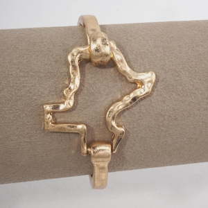 May include: A gold-toned metal bracelet featuring a central, irregular, organic-shaped link with a textured, hammered surface. The design includes a hinged clasp mechanism that connects the wavy, abstract frame, creating a continuous loop structure.
