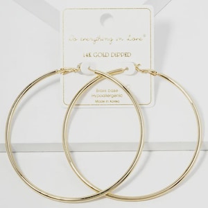 May include: A pair of large, thin, gold-toned hoop earrings attached to a white display card. The card features the text Do everything in love, 14K GOLD DIPPED, Brass base, Hypoallergenic, and Made in Korea printed in black and gold ink.