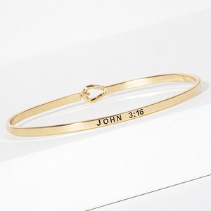 May include: A gold-toned metal bangle bracelet featuring a smooth, polished finish and a hook-and-eye style closure. The exterior surface of the bracelet is engraved with the black text "JOHN 3:16" in a clean, sans-serif font style.