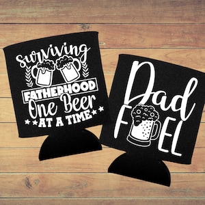 Op de afbeelding: Twee zwarte blikjeskoelers met witte tekst en afbeeldingen. De eerste koeler zegt "Surviving Fatherhood One Beer At A Time" met twee bierglazen en tarwehalmen. De tweede koeler zegt "Dad Fuel" met een bierglas.
