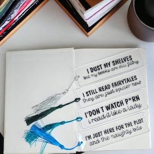 May include: Four clear acrylic bookmarks with black text. The bookmarks have funny sayings about reading and books. The bookmarks are on a white surface with a book and a mug in the background.
