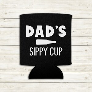 Op de afbeelding: Een zwarte neopreen blikjeskoeler met witte tekst die "DAD'S SIPPY CUP" zegt en een wit silhouet van een bierfles.