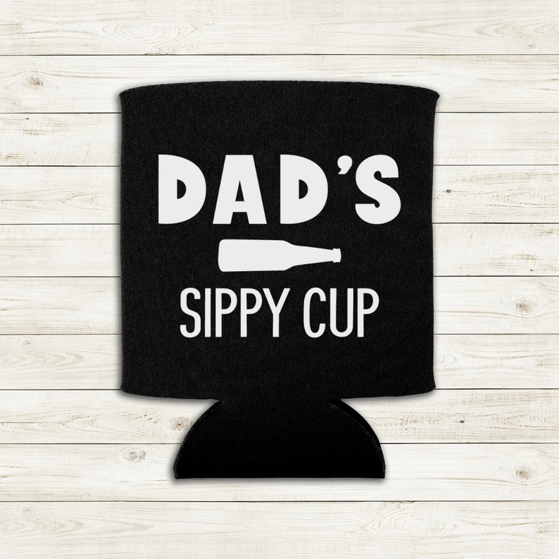 Op de afbeelding: Een zwarte neopreen blikjeskoeler met witte tekst die "DAD'S SIPPY CUP" zegt en een wit silhouet van een bierfles.