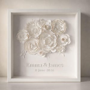 May include: A white framed paper flower art piece. The artwork features a bouquet of various paper flowers in shades of white. The names "Emma & James" and the date "11 June 2026" are embossed on the bottom.