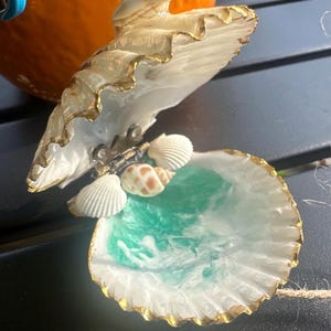 May include: An open seashell ornament with gold-painted edges, featuring a hinged metal mechanism. The interior is filled with a turquoise and white resin wave design, accented by three small, natural seashells attached near the hinge.
