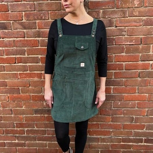 May include: A woman wearing a green corduroy pinafore dress with black leggings and black trainers. She is standing in front of a brick wall.
