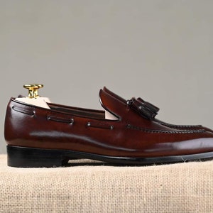 Puede incluir: Un par de mocasines de cuero marr&oacute;n con detalles de borlas. Los zapatos tienen un dise&ntilde;o cl&aacute;sico con un tac&oacute;n bajo y se exhiben sobre un fondo neutro. El cuero parece pulido y las costuras son visibles.
