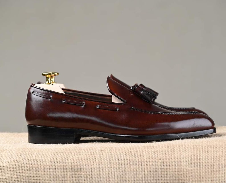 Puede incluir: Un par de mocasines de cuero marr&oacute;n con detalles de borlas. Los zapatos tienen un dise&ntilde;o cl&aacute;sico con un tac&oacute;n bajo y se exhiben sobre un fondo neutro. El cuero parece pulido y las costuras son visibles.