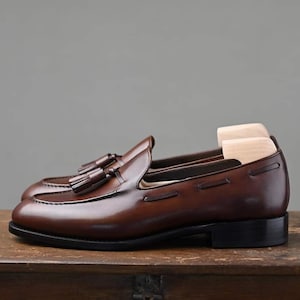Puede incluir: Un par de mocasines de cuero marr&oacute;n con detalles de borlas. Los zapatos tienen un acabado pulido y una suela negra. Se insertan hormas de madera. Los mocasines se exhiben sobre una superficie de madera.