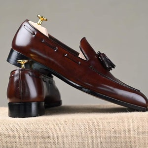 Puede incluir: Un par de mocasines de cuero marr&oacute;n oscuro y pulido con detalles de borlas. Los zapatos se exhiben sobre una superficie neutra, mostrando su dise&ntilde;o elegante y artesan&iacute;a. Se insertan hormas para mantener la forma.