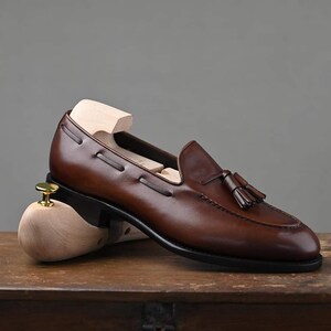 Puede incluir: Mocas&iacute;n de cuero marr&oacute;n con detalle de borlas. El zapato tiene un dise&ntilde;o cl&aacute;sico con punta redondeada y suela de cuero. Se inserta una horma de madera dentro del zapato. El mocas&iacute;n se exhibe sobre una superficie de madera.