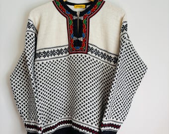 Könnte beinhalten: Ein weißer Strickpullover mit einem schwarz-weißen geometrischen Muster. Der Pullover hat einen bunten bestickten Ausschnitt mit einem dekorativen Metallverschluss.
