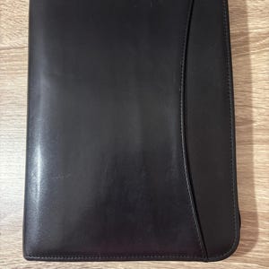 May include: A black leather portfolio with a curved design on the right side. The portfolio is closed and appears to be made of a smooth, possibly polished, leather material. The background is a light brown wooden surface.