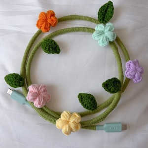 May include: A decorative USB-C charging cable with a green, vine-like design, embellished with crocheted flowers in orange, pink, yellow, purple, and light blue. The cable also features green crocheted leaves. The cable ends are a light blue colour.